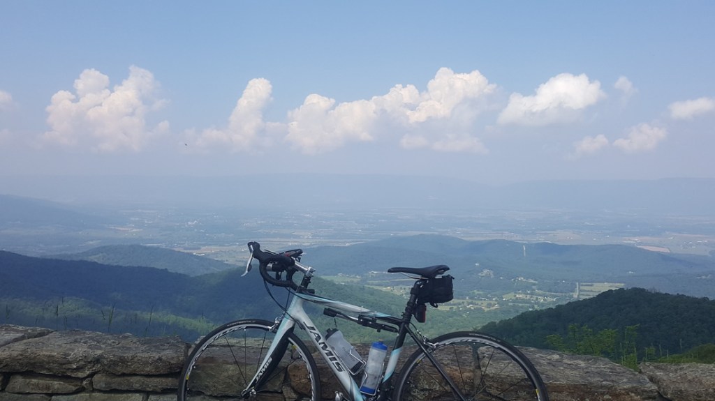 Natalie's bike leans up against a stone wall overlooking a valley with rolling tree covered hills and a few small populated areas. The sky is blue with fluffy white clouds.